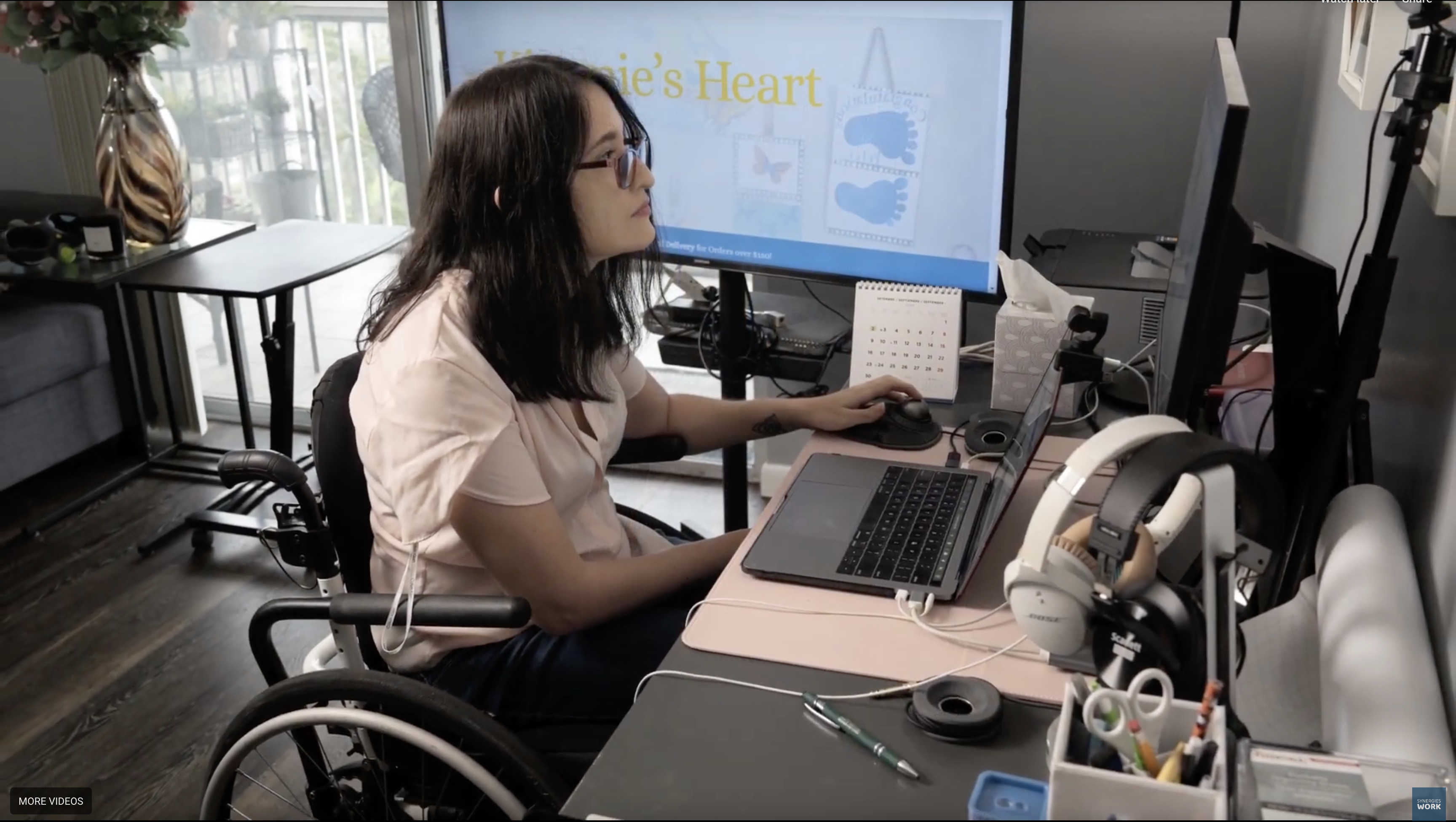 A woman in a wheelchair sits at a desk with a laptop, wearing sunglasses, focused on work. A screen displays "Erica's Heart."